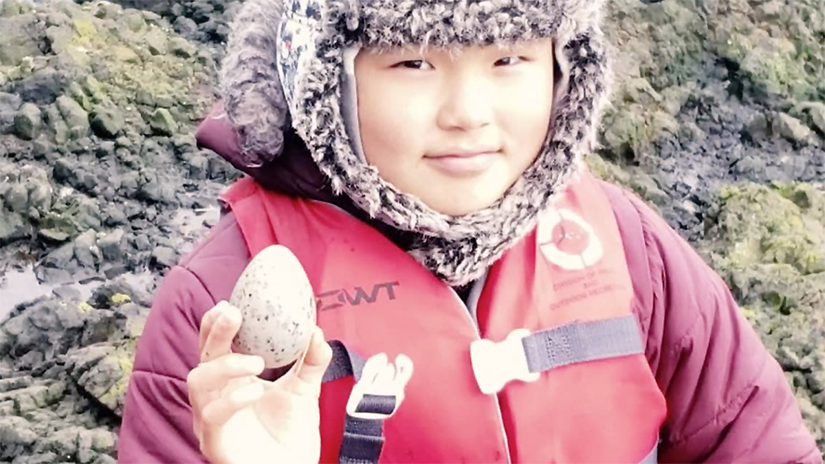 Healthy Tribes TPWIC Alaskan Boy with Egg Native Alaskan boy in winter coat and life vest on rocky shore holding up a bird's egg.