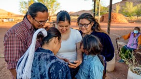 NCCDPHPInvestSDOH A group of five people in a rural environment standing in a circle looking at a phone together.